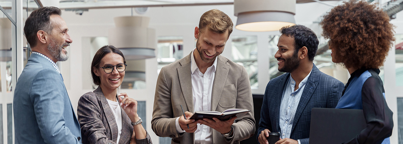 Ein dynamisches Team von f&uuml;nf Gesch&auml;ftsleuten tauscht sich in einer modernen B&uuml;roumgebung aus. Die lockere und freundliche Atmosph&auml;re, begleitet von Lachen und positiver Energie, unterstreicht die Bedeutung von Teamarbeit, Kommunikation und beruflicher Entwicklung.