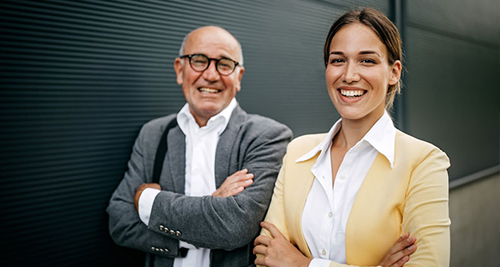 Zwei l&auml;chelnde Personen, ein &auml;lterer Mann mit Brille und grauem Sakko und eine j&uuml;ngere Frau mit gelbem Blazer, stehen vor einer dunklen Wand mit verschr&auml;nkten Armen.