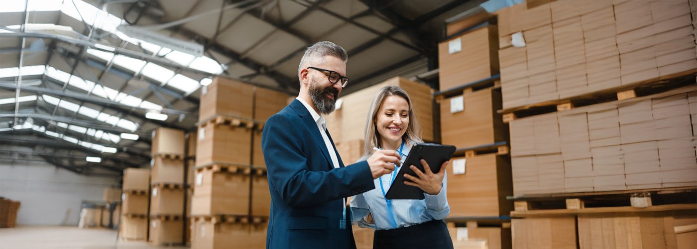 A man and a woman in a warehouse with packed, stacked pallets in the background. The woman is holding a clipboard, the man a pen. Both are looking at the clipboard.