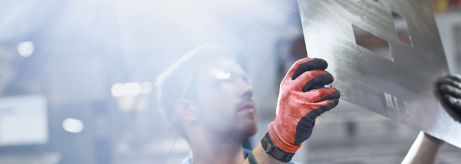 A man wearing red protective gloves holds a metal plate in a production hall and examines it.